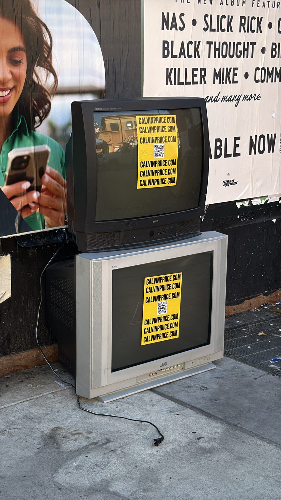 Yellow CALVINPRIICE.COM stickers on two stacked CRT TVs dumped on an LA sidewalk, in front of a major-label hip-hop billboard