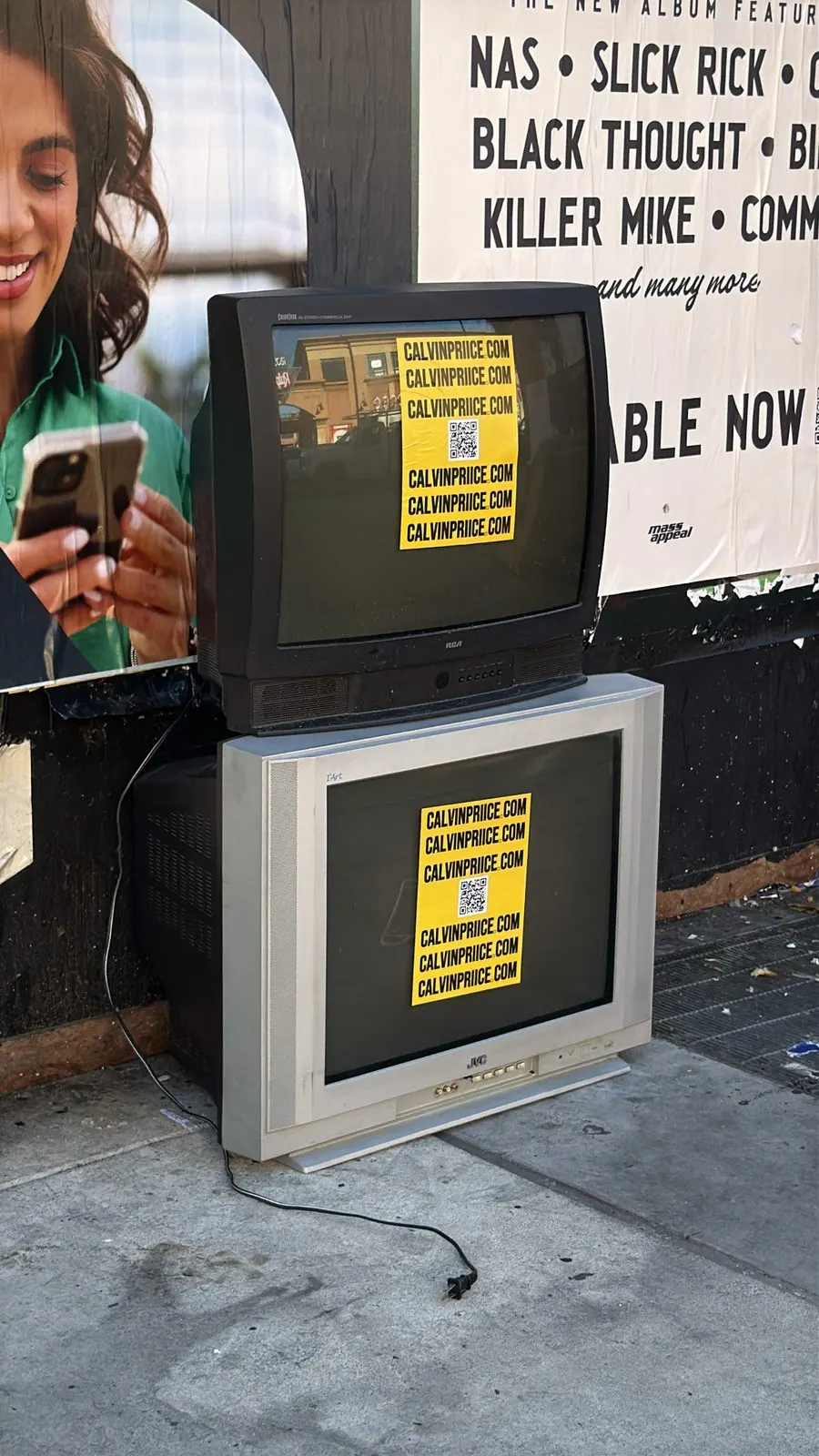 Yellow CALVINPRIICE.COM stickers on two stacked CRT TVs dumped on an LA sidewalk, in front of a major-label hip-hop billboard